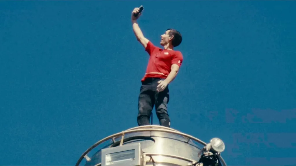 艾力克斯．霍諾德站在台北101高處，手持手機自拍，背景為晴朗藍天。