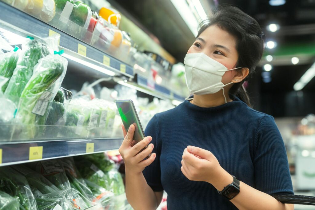 Young adult asian female woman wearing protective face mask hand choosing fresh product consumer product groceries store department mall with care cheerful new normal shopping lifestyle